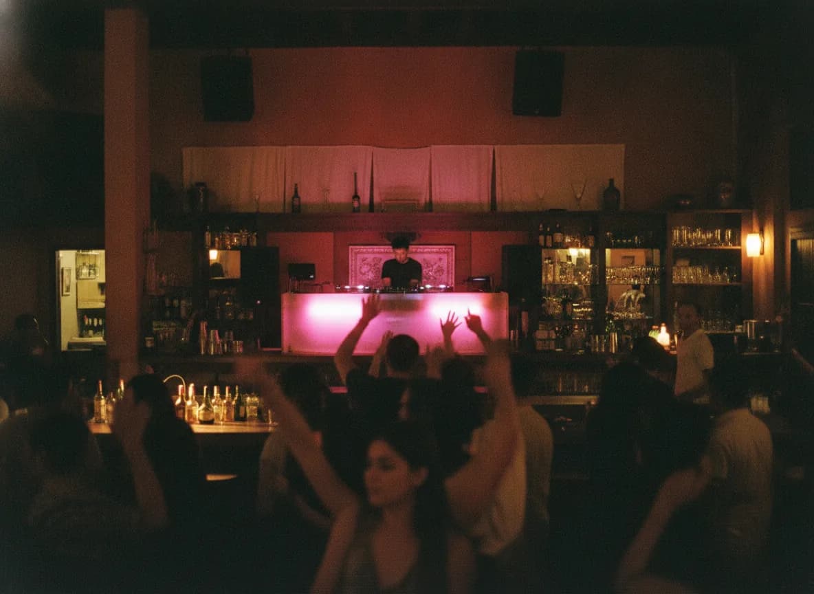 Bar de noche con la gente bailando frente a la cabina del DJ, iluminada de rosa